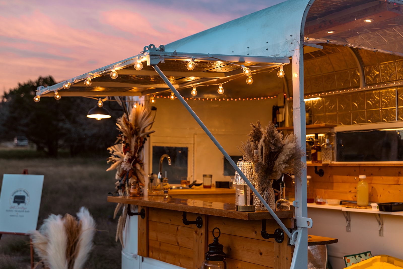 Unbridled Trailers mobile bar lit up at dusk with string lights and pampas grass